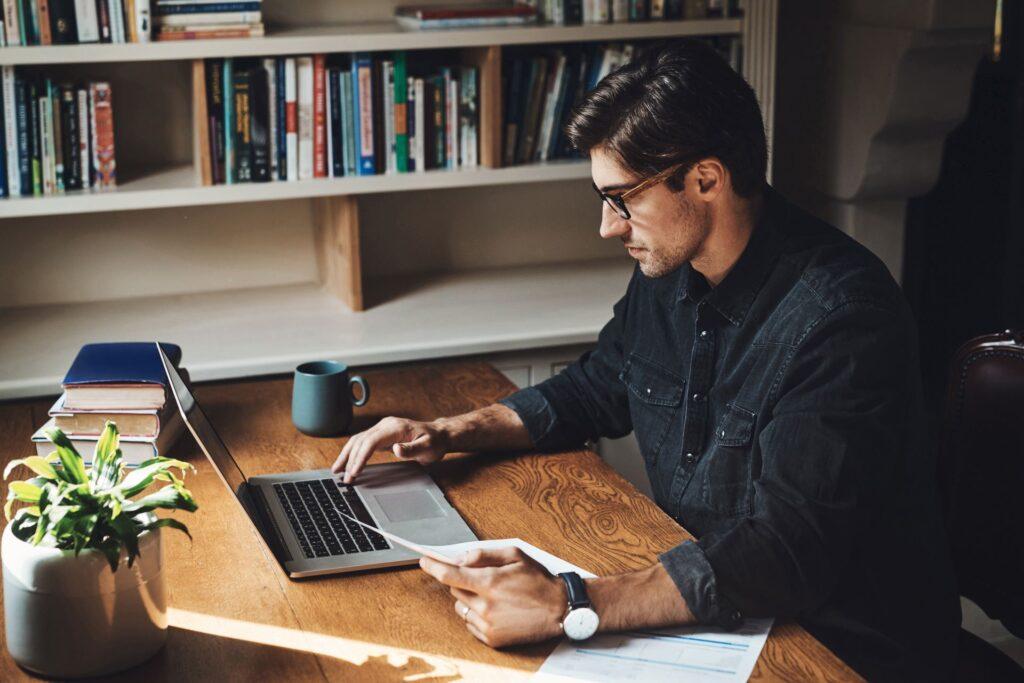 Ein Mann arbeitet konzentriert im Homeoffice an seinem Laptop, umgeben von Büchern und Dokumenten. Das Bild zeigt die sorgfältige Vorbereitung auf eine Steuerberatung zur Optimierung der Finanzen.